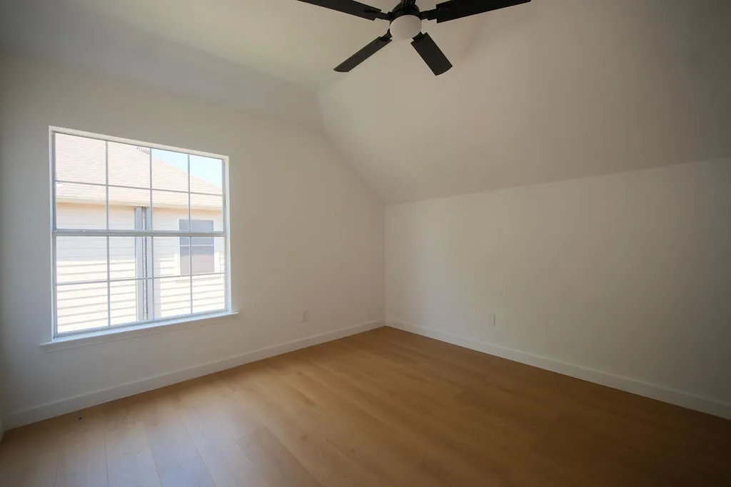 Bright corner bedroom featuring abundant natural light from large multi-pane windows, gleaming wood-finish flooring throughout, vaulted ceiling with integrated ceiling fan, and crisp white walls providing a clean, neutral backdrop