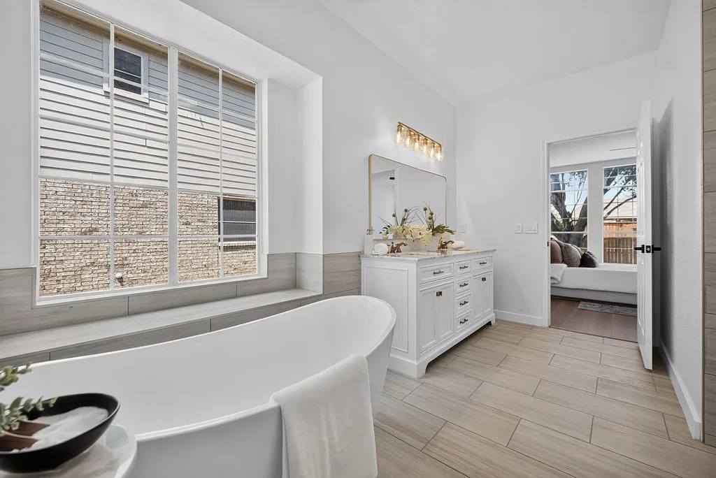 Spa-inspired primary bathroom featuring a freestanding soaking tub, white cabinetry with multiple drawers, wood-finish flooring in herringbone pattern, and abundant natural light from large windows with plantation shutters