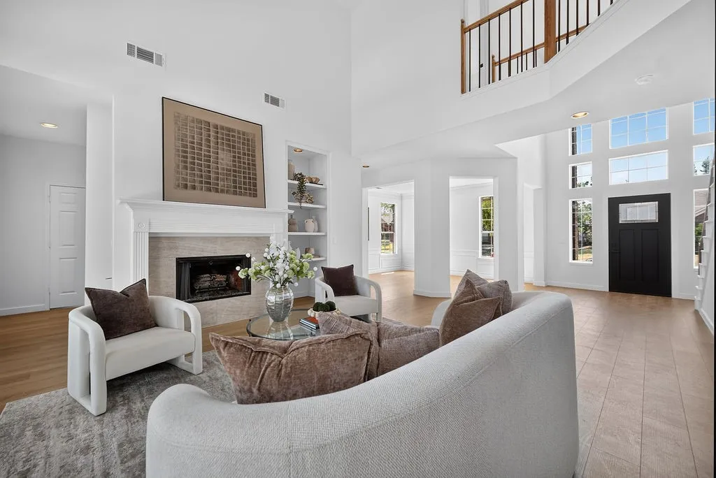Bright, open-concept living area featuring a two-story ceiling with skylights, a wood-burning fireplace with white surround and built-in shelving, hardwood flooring, and abundant natural light from multiple windows and glass doors