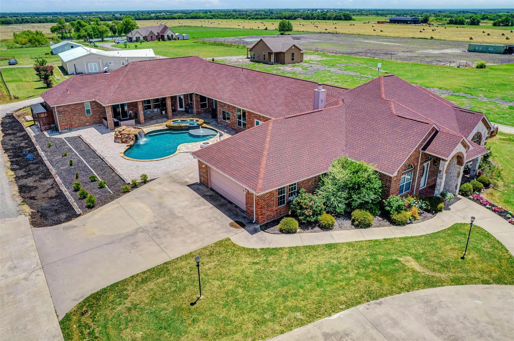 Sprawling brick residence with distinctive burgundy metal roof and curved architectural lines, anchored by an in-ground swimming pool with integrated spa