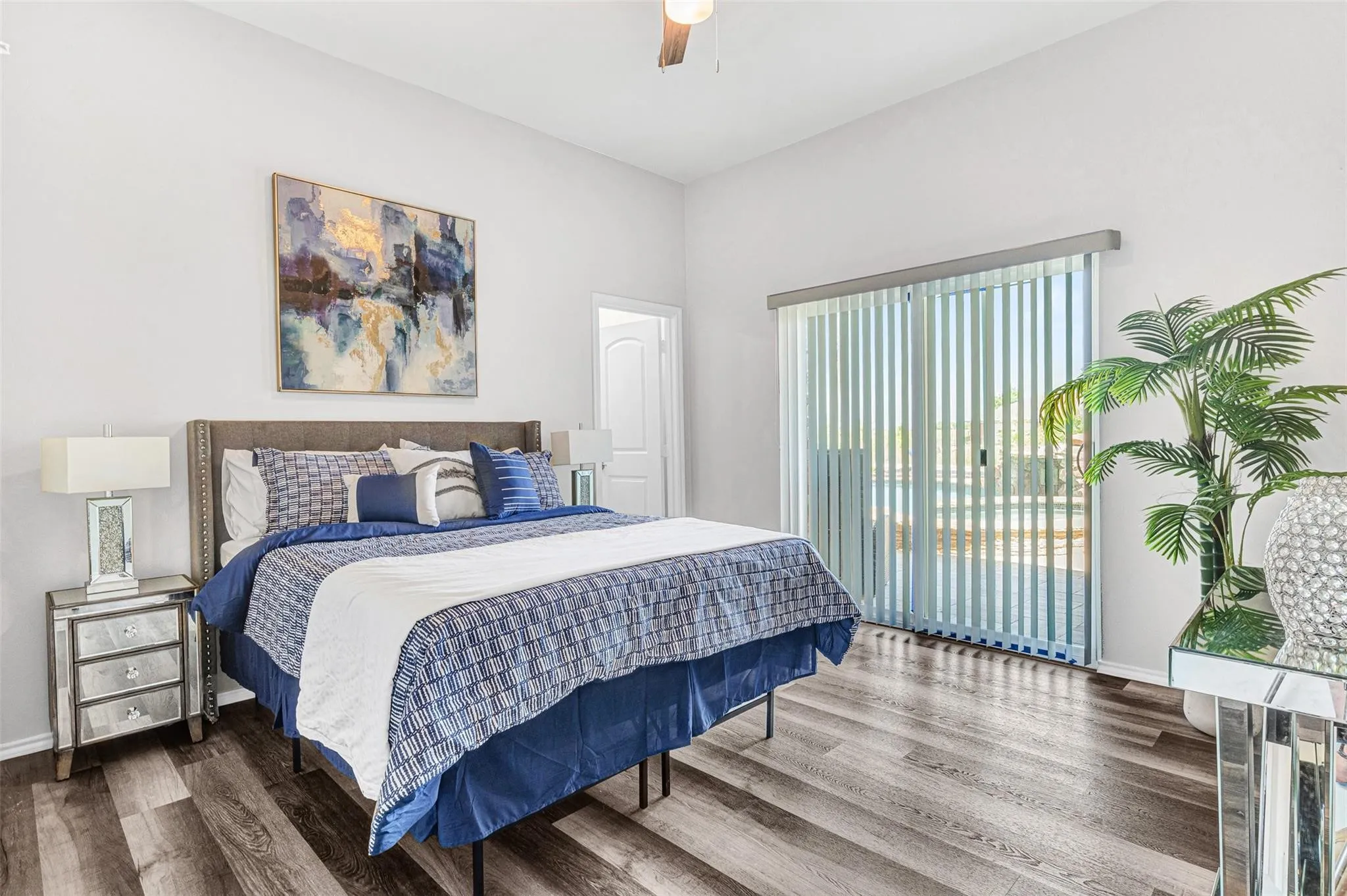 Spacious primary bedroom showcasing a wood-finish plank floor, soft-close dresser with multiple drawers, bedside table with integrated lighting, large sliding glass door with vertical blinds providing natural light, and a vibrant abstract artwork above the headboard