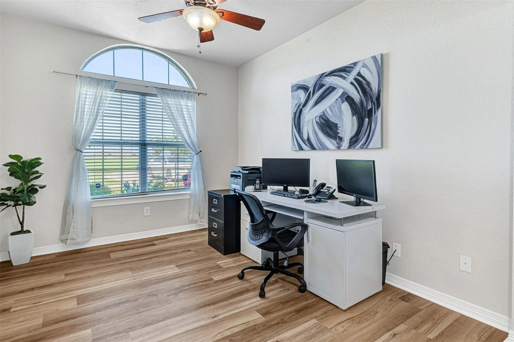 Bright home office featuring hardwood flooring, a curved arched window with plantation shutters and natural light, dual monitor setup with built-in desk cabinetry, an ergonomic office chair, ceiling fan with integrated lighting, and abstract wall art
