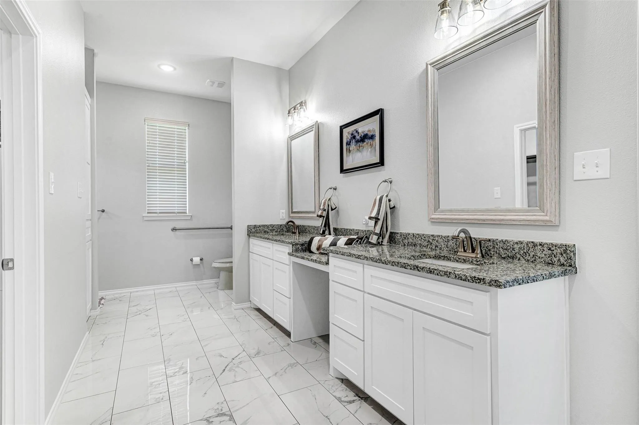 Spa-inspired primary bathroom featuring dual vanities with white cabinetry, granite countertops, polished chrome fixtures, and a large framed mirror