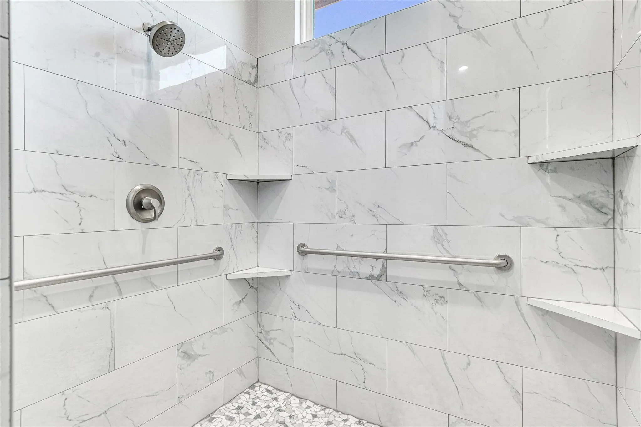 Spa-inspired shower enclosure featuring large-format marble-look tile walls, a rainfall showerhead, dual wall-mounted grab bars, a built-in corner bench seat, and pebble flooring detail