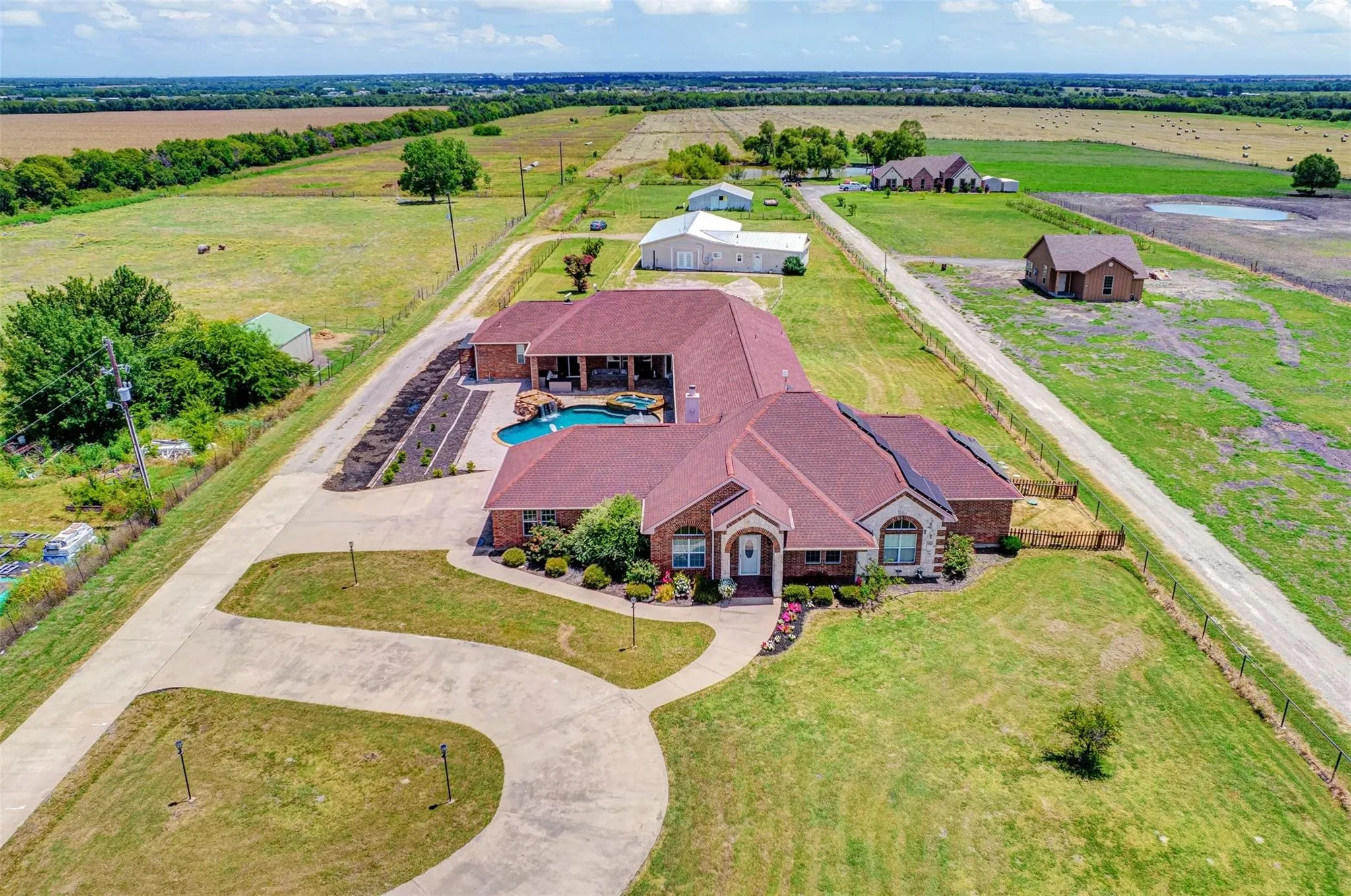 Brick residence with a burgundy metal roof situated on expansive acreage, featuring a curved driveway approach, manicured lawn, mature landscaping, and detached outbuildings visible across the​​‌​​​​‌​​‌‌​‌‌​​​‌‌​‌​‌​‌​​​‌​​ property