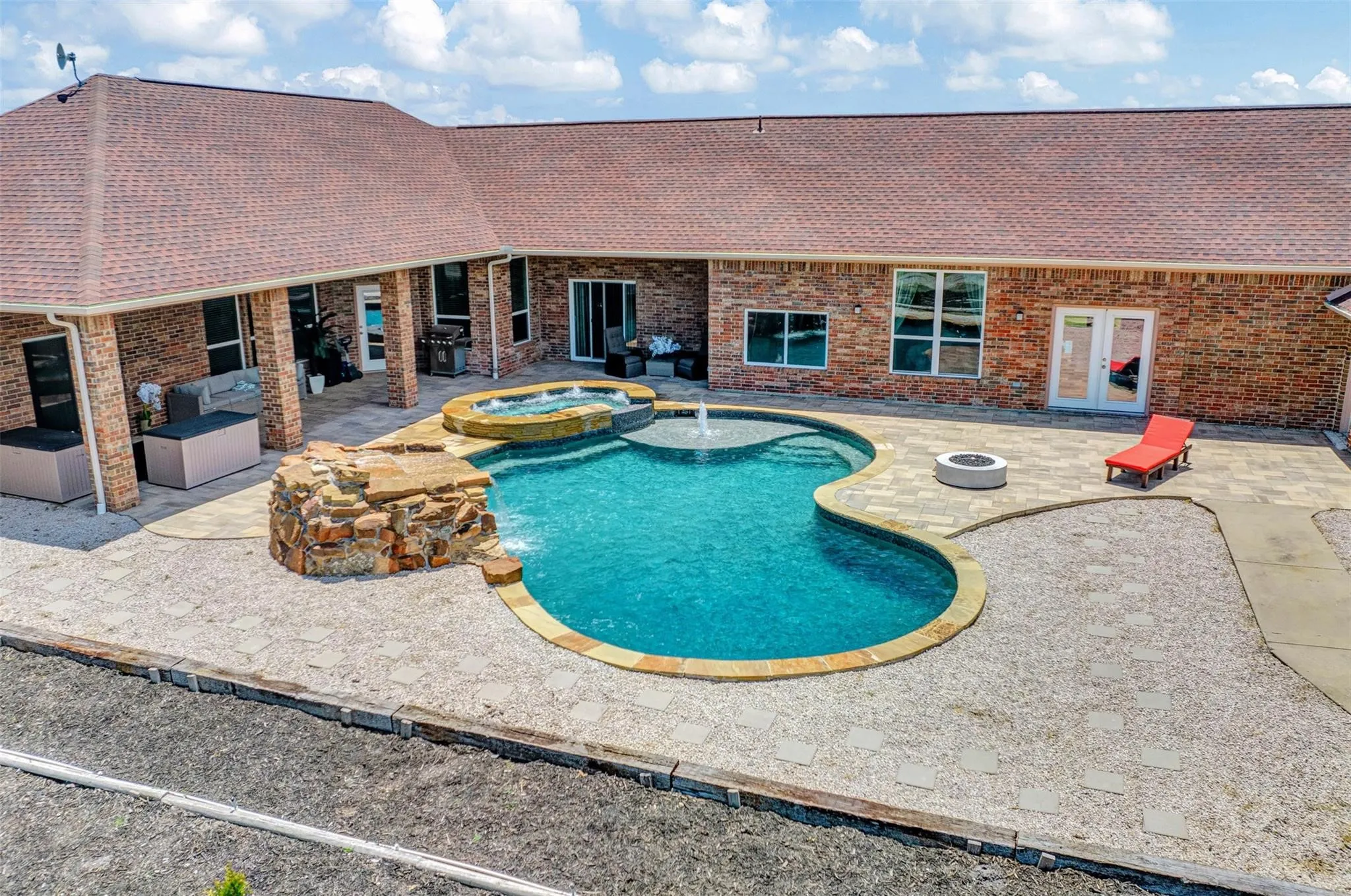 Expansive backyard oasis featuring a custom free-form swimming pool with integrated spa, decorative stone water feature, concrete decking, and brick construction home with multiple glass door access points and covered patio areas overlooking the resort-style outdoor entertaining space