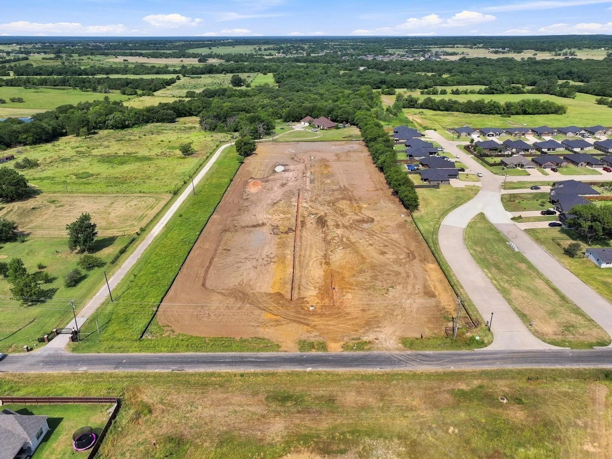 Cleared lot with direct road access, adjacent to developed residential areas, surrounded by green fields and distant trees
