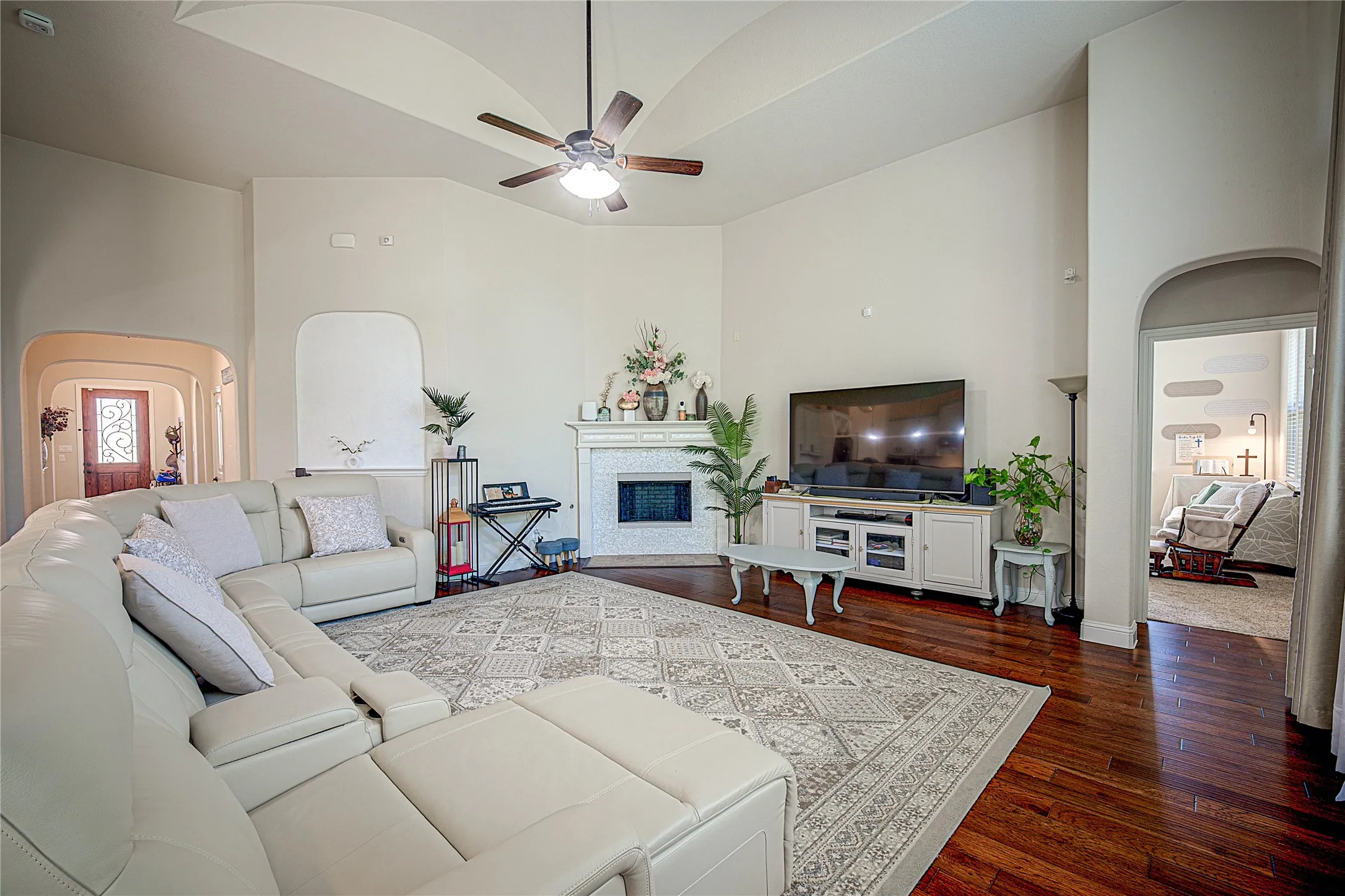 Vaulted ceilings with a ceiling fan, hardwood flooring, fireplace with white mantle, arched doorways, and built-in shelving