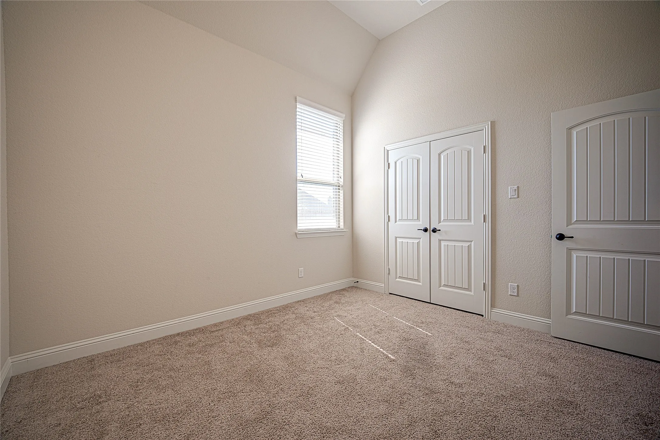 Neutral-toned carpeting, vaulted ceiling, single window with blinds, white paneled bifold doors, and a white paneled door with matte black hardware
