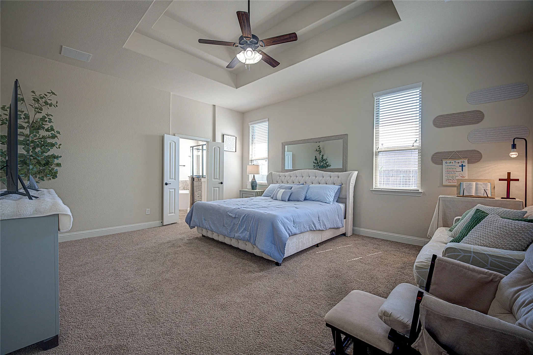Tray ceiling with a ceiling fan, two windows with blinds, and double doors opening to a bathroom