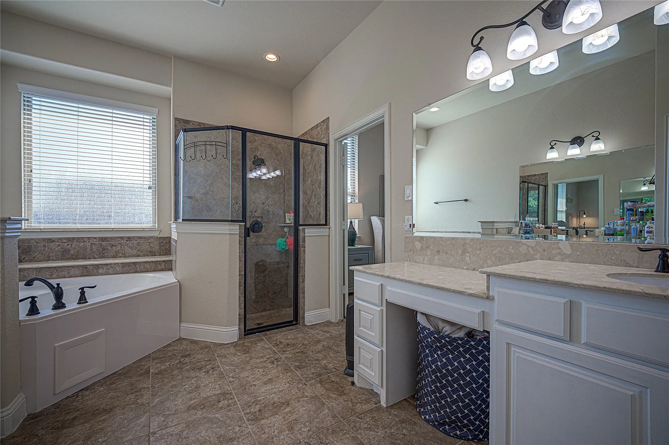 Corner garden tub with dark bronze fixtures, walk-in shower with glass enclosure, dual vanity with light-toned countertops, and large vanity mirror with overhead lighting