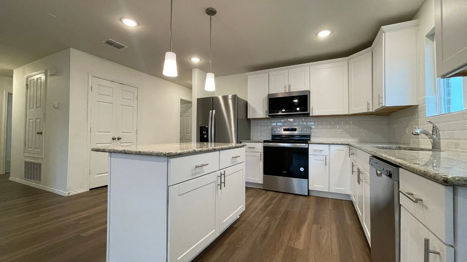 Modern kitchen featuring white cabinetry, stainless steel appliances including a double wall oven and refrigerator, quartz countertops, a centered island with seating space, pendant lighting, subway tile backsplash, and wood-finish flooring throughout the open layout