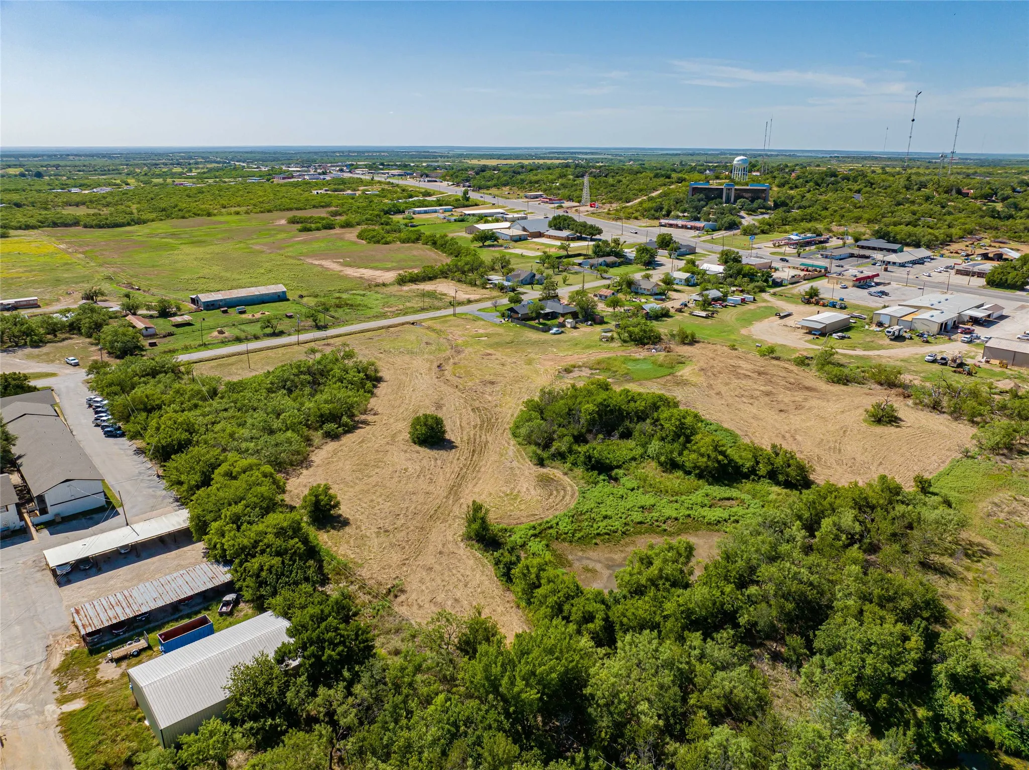 Expansive aerial view showcasing approximately 20+ acres of mixed-use property featuring cleared land, mature tree clusters, and established infrastructure including multiple commercial buildings, paved parking areas, and utility access across the grounds