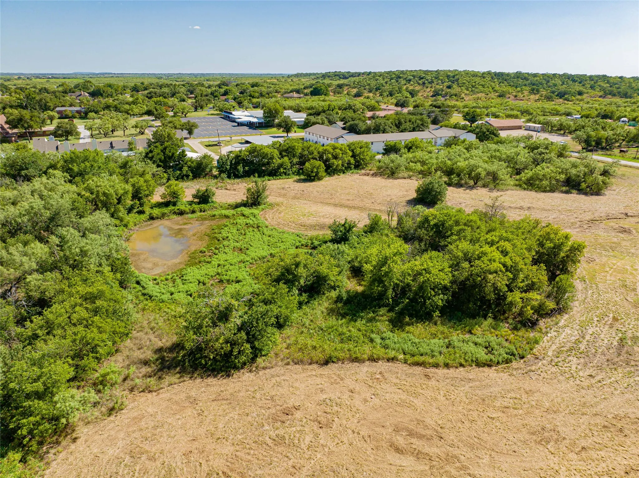 Expansive rural land featuring natural vegetation, a tranquil pond, and distant residential structures