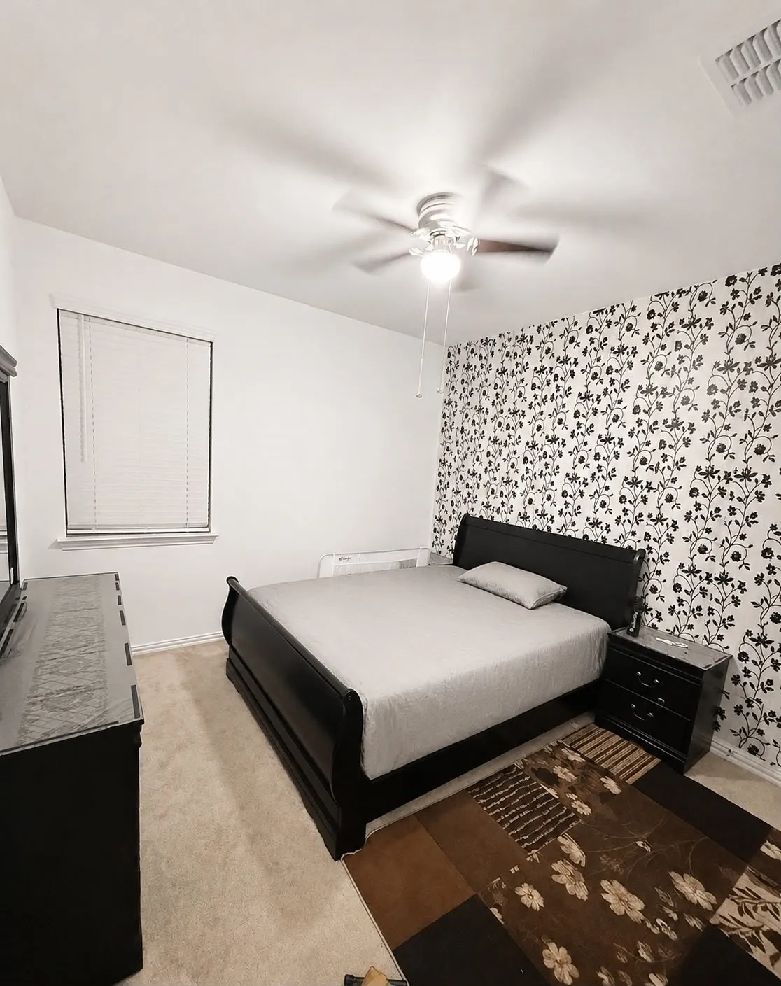 Spacious bedroom featuring a black platform bed frame, wood-finish flooring, ceiling fan with integrated lighting, patterned accent wall, and a large window with natural light