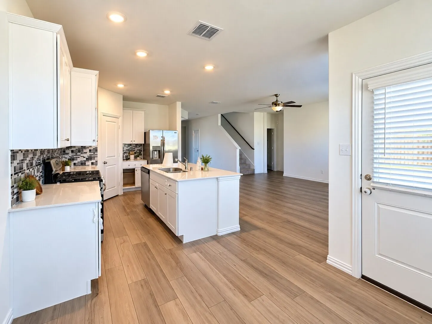 Modern kitchen featuring white cabinetry, a central island with an integrated sink, mosaic tile backsplash, stainless steel appliances, and wood-finish flooring
