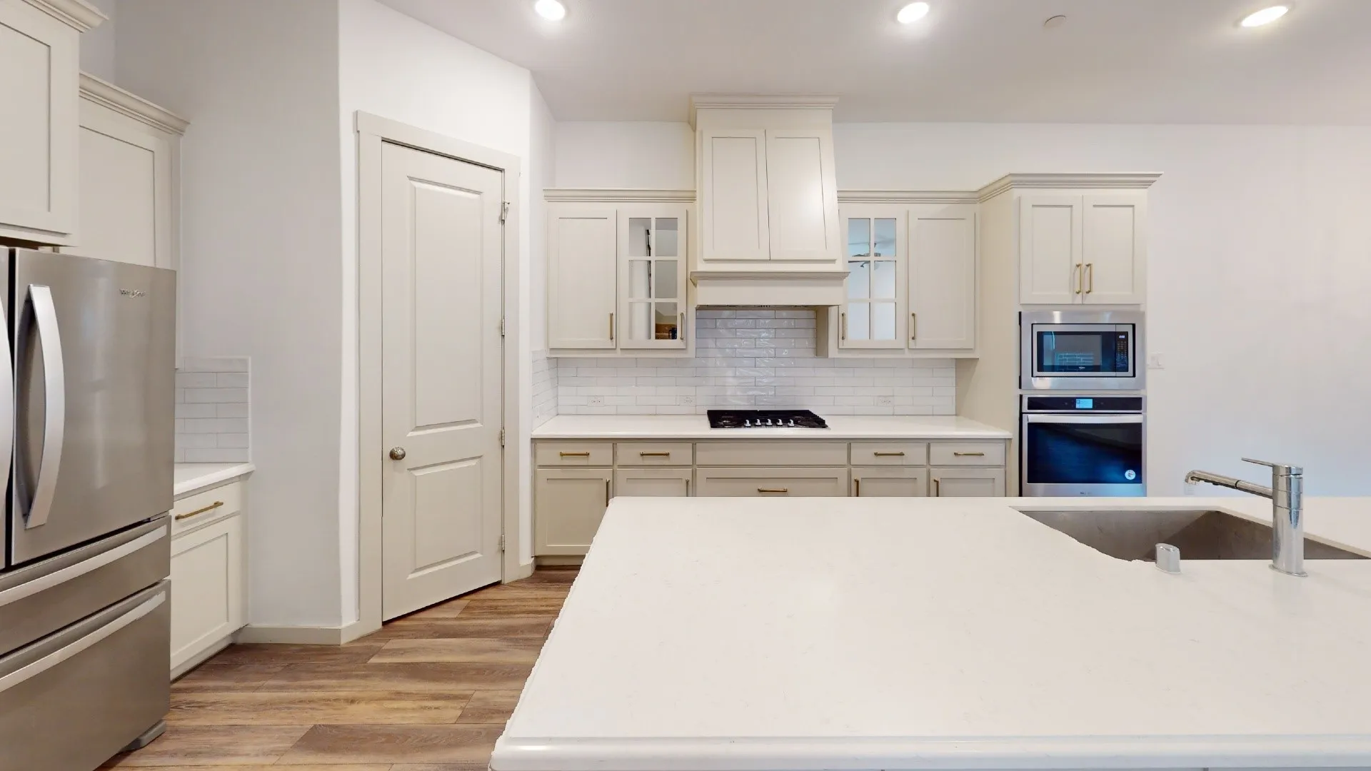 Spacious kitchen featuring an integrated range hood, stainless steel appliances, white subway tile backsplash, light wood-finish flooring, and a large island with an undermount sink