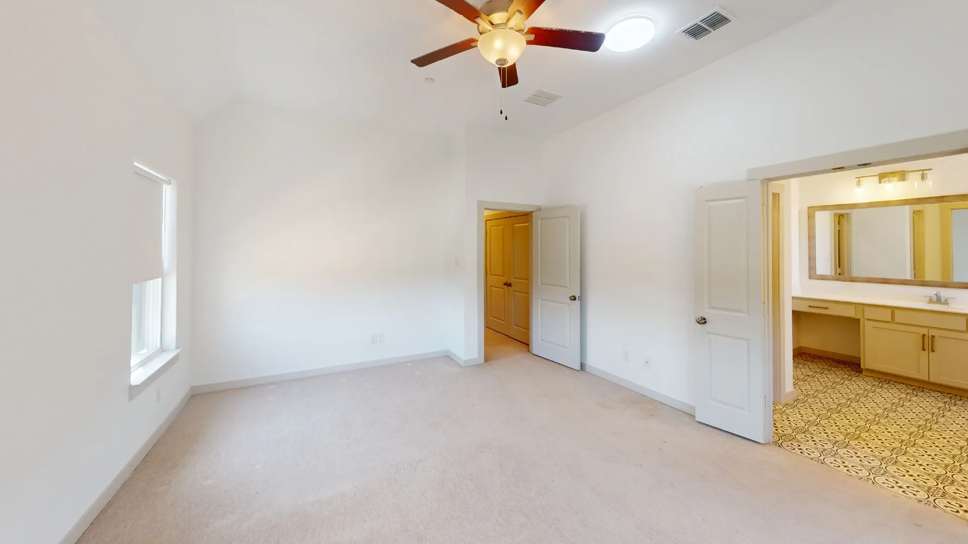 Spacious primary bedroom suite featuring neutral carpeting, ceiling fan with integrated lighting, and direct access to an ensuite bathroom with dual vanity and abundant storage cabinetry