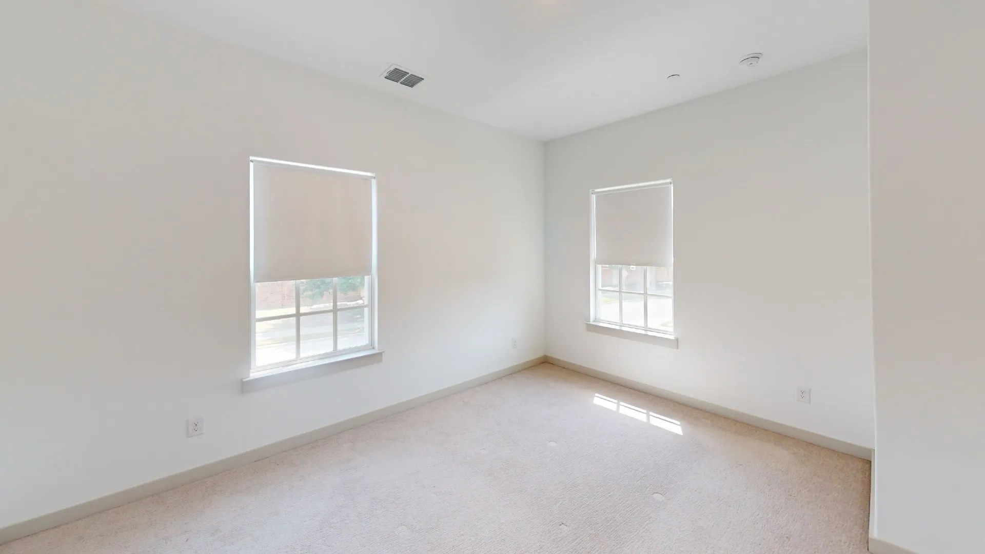 Bright secondary bedroom with dual windows featuring roller shades, neutral wall finishes, and neutral-toned wood-finish flooring throughout