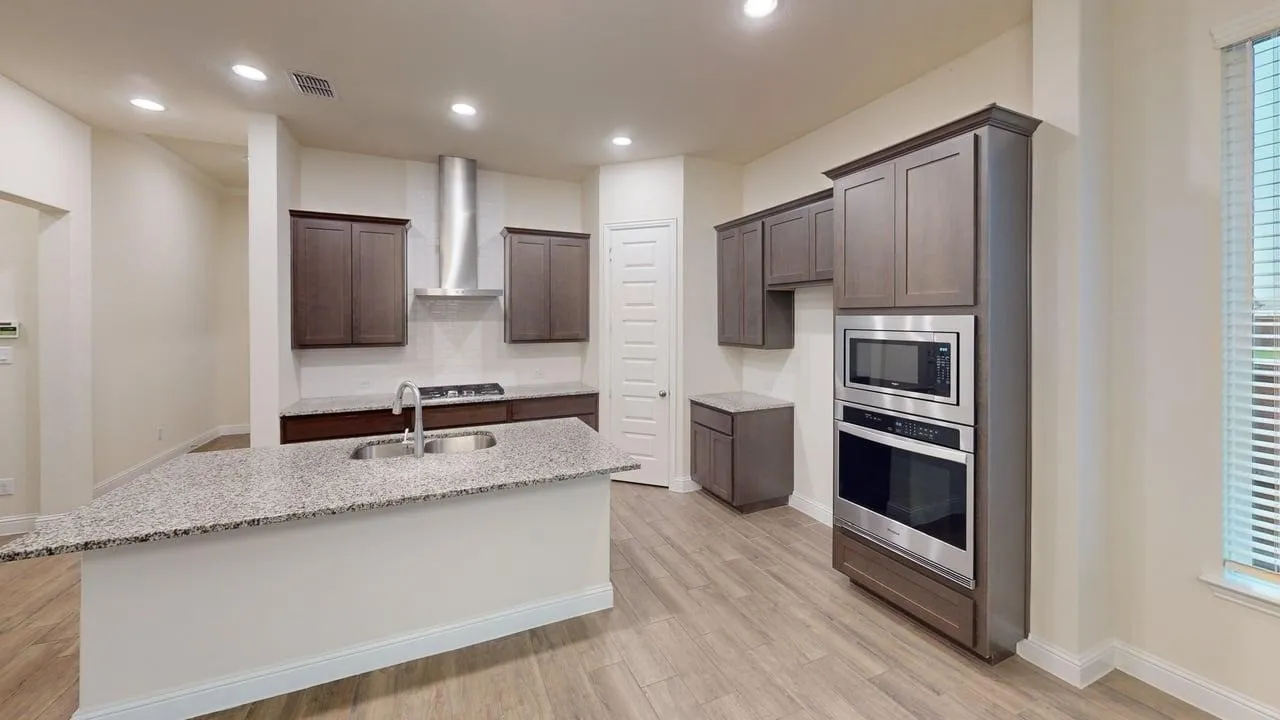 Modern kitchen featuring a large center island with waterfall-edge countertop, stainless steel appliances including dual wall ovens and a gas cooktop with overhead hood, dark cabinetry, granite countertops, wood-finish flooring, and abundant recessed lighting throughout the open layout