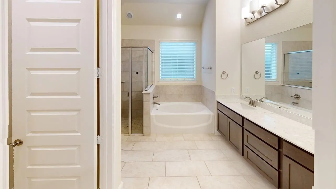Spa-inspired primary bathroom featuring a soaking tub with glass enclosure, dual-sink vanity with wood cabinetry and quartz countertop, large framed mirrors with integrated lighting, tile flooring, and a window with plantation shutters