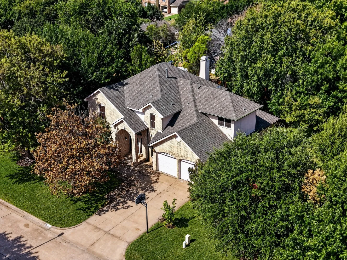 Traditional style residence featuring a light brick exterior, an arched entry, a two-car garage, mature trees, and a shingled roof - virtually added green grass