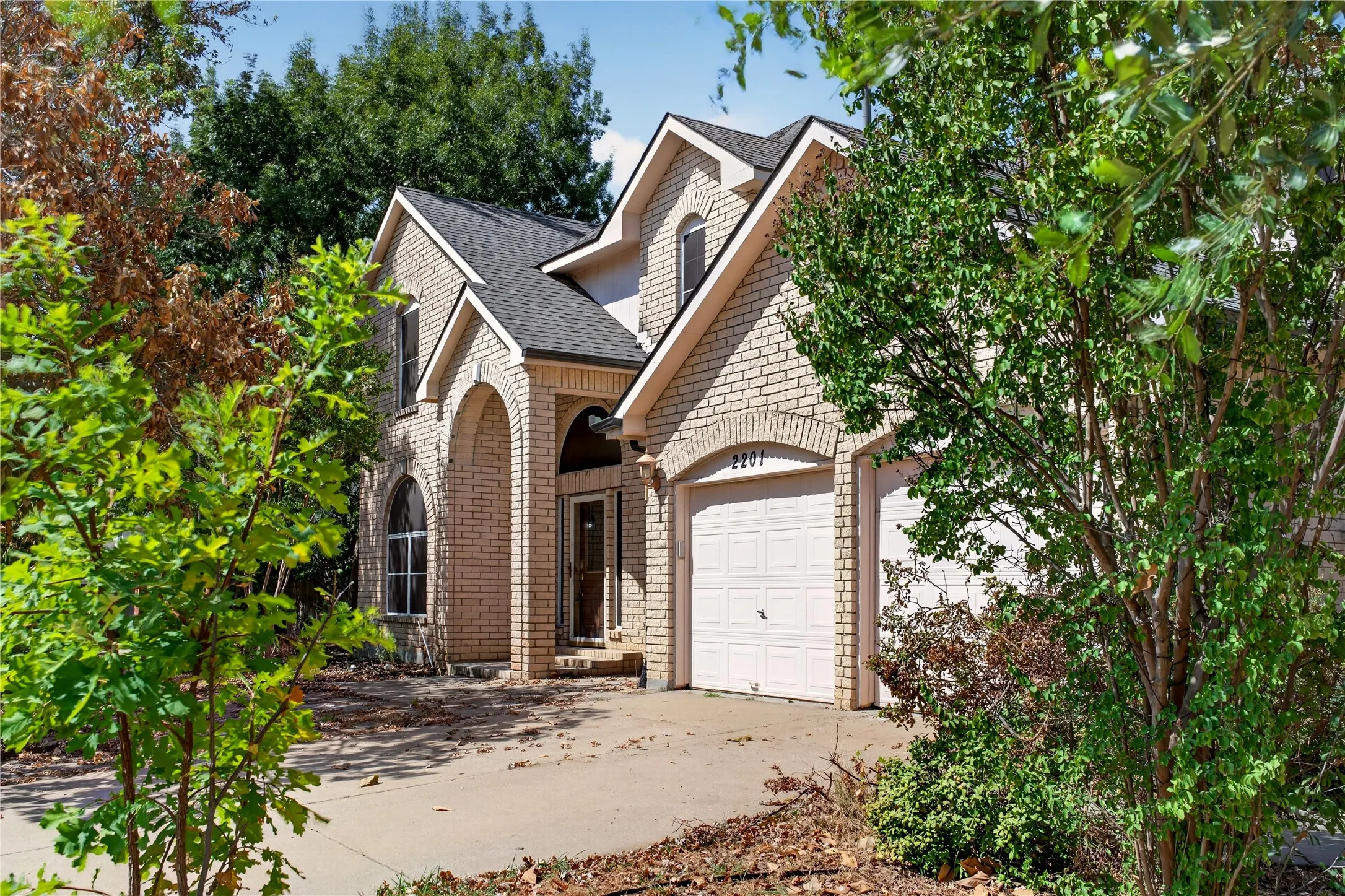 Brick exterior with a gabled roofline, arched entry, two-car garage, and established trees