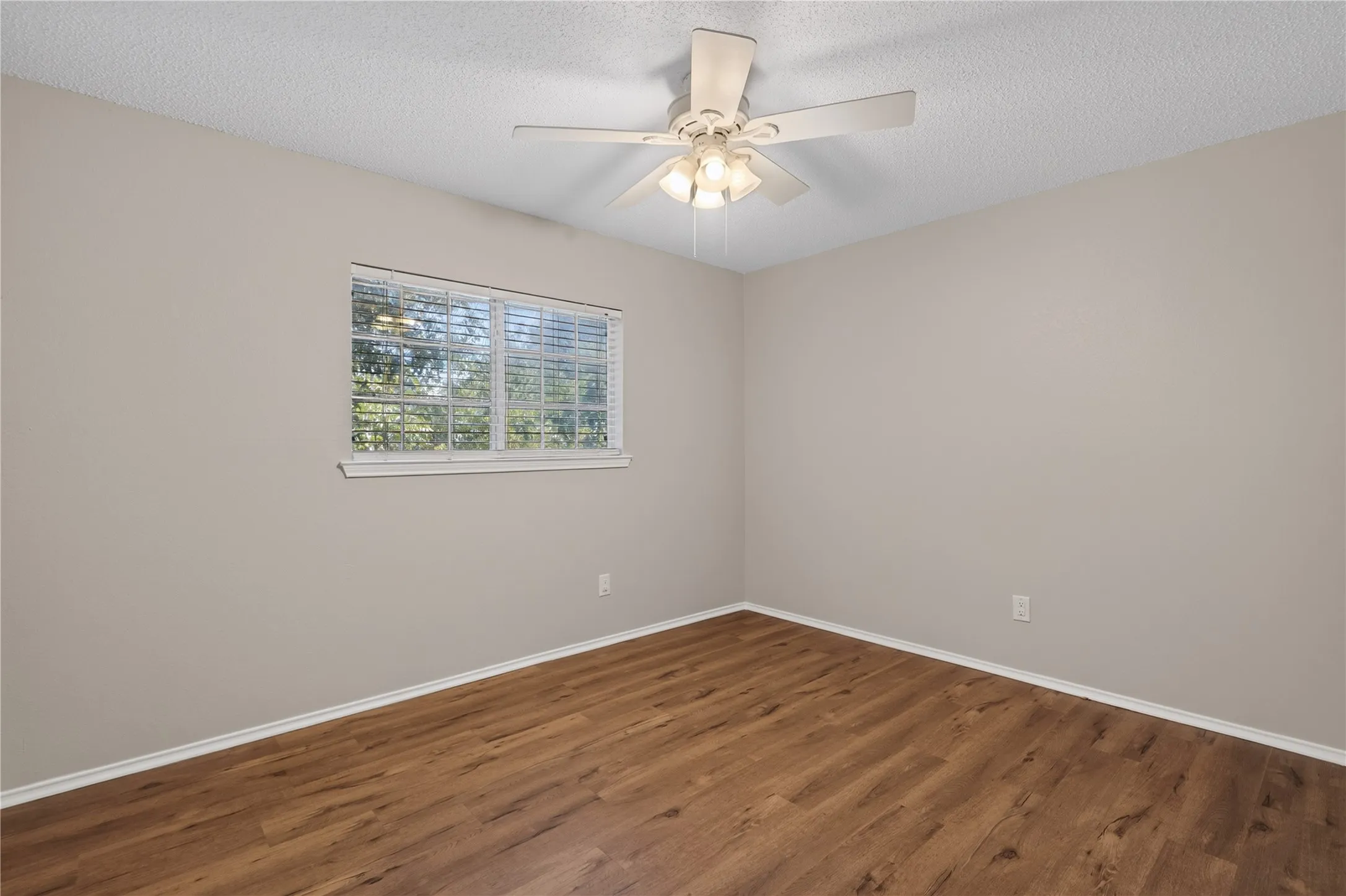 Wood-finish flooring with white trim, a single window with blinds, and an overhead ceiling fan with integrated lighting