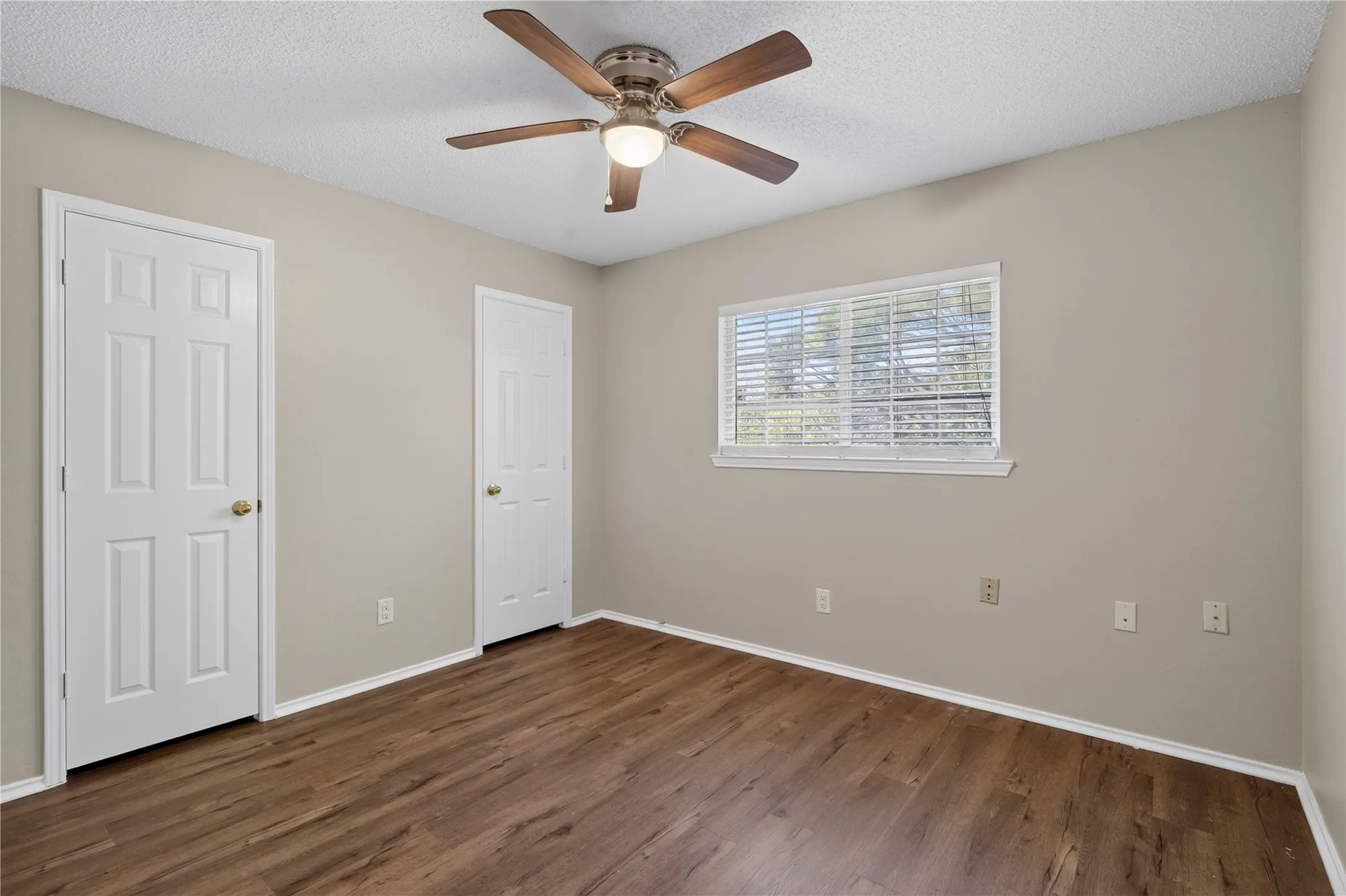 Wood-finish flooring, neutral wall tones, window with horizontal blinds, and a ceiling fan with integrated lighting