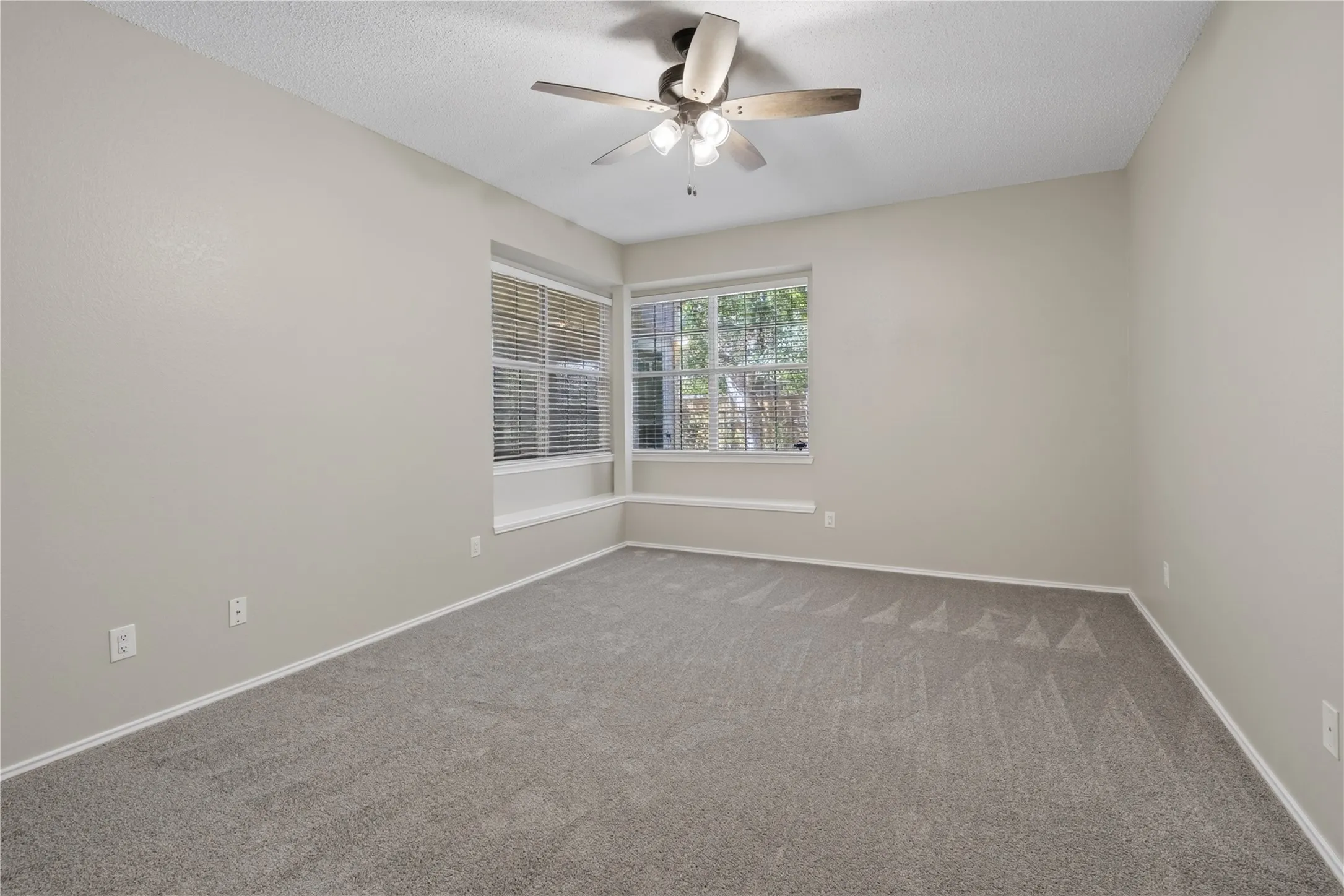 Neutral-toned room featuring a ceiling fan with integrated lighting, multiple windows with horizontal blinds, and grey patterned carpeting