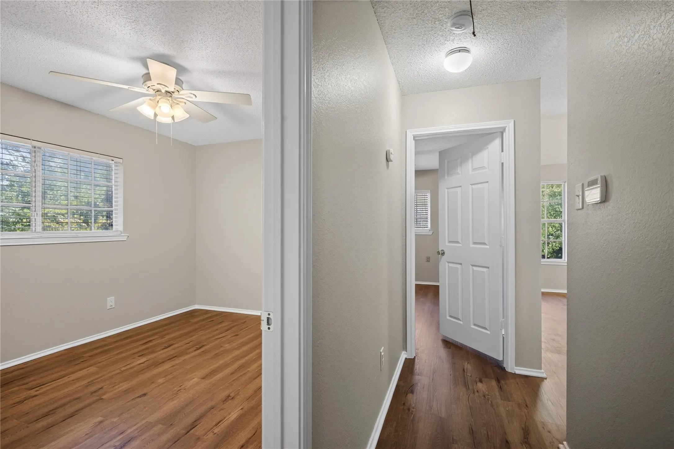 Wood-finish flooring throughout, ceiling fan with light fixture, window with blinds, paneled interior doors, and a wall-mounted thermostat