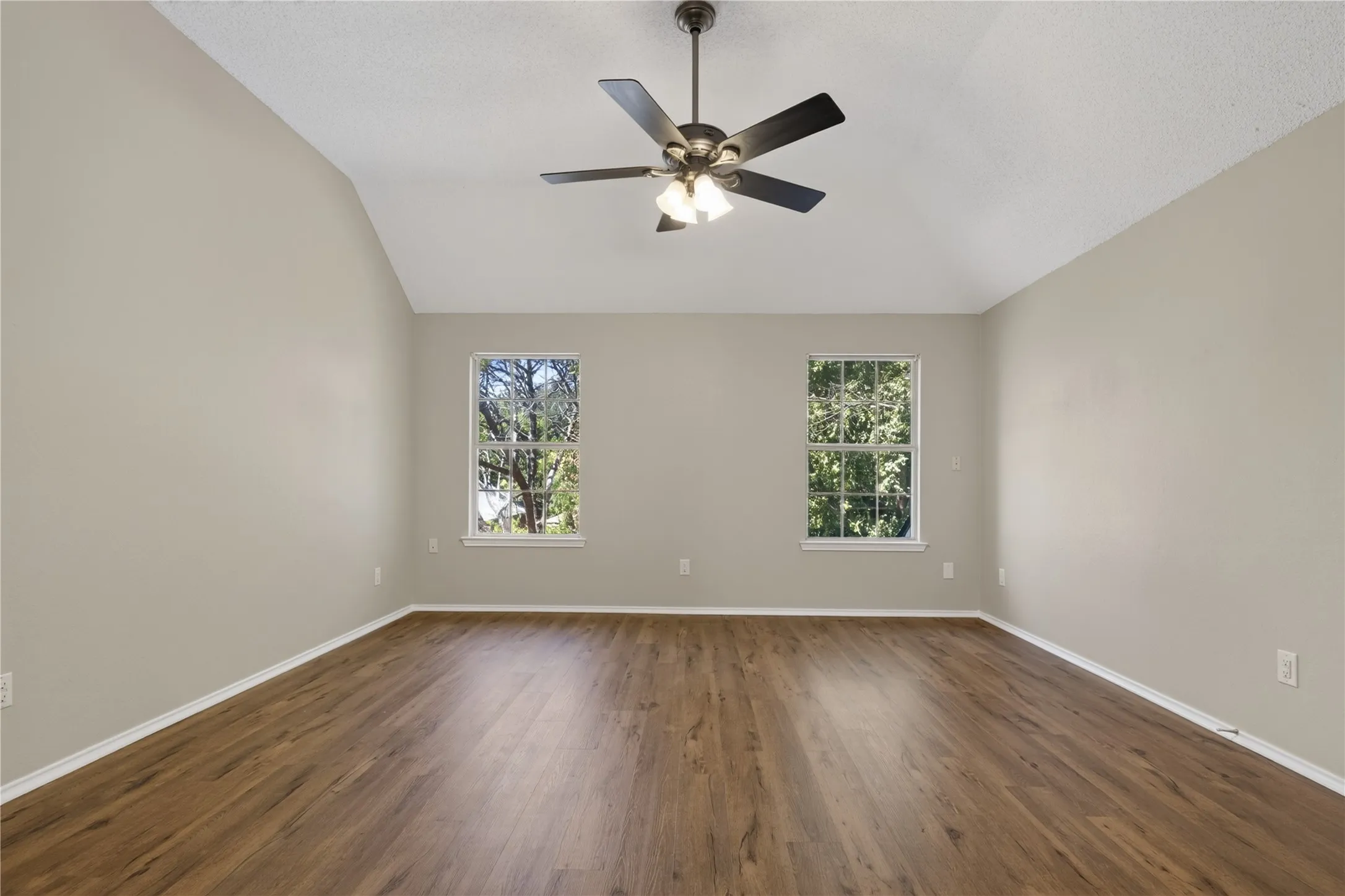 Vaulted ceiling with a ceiling fan, two windows, wood-finish flooring, and neutral wall tones