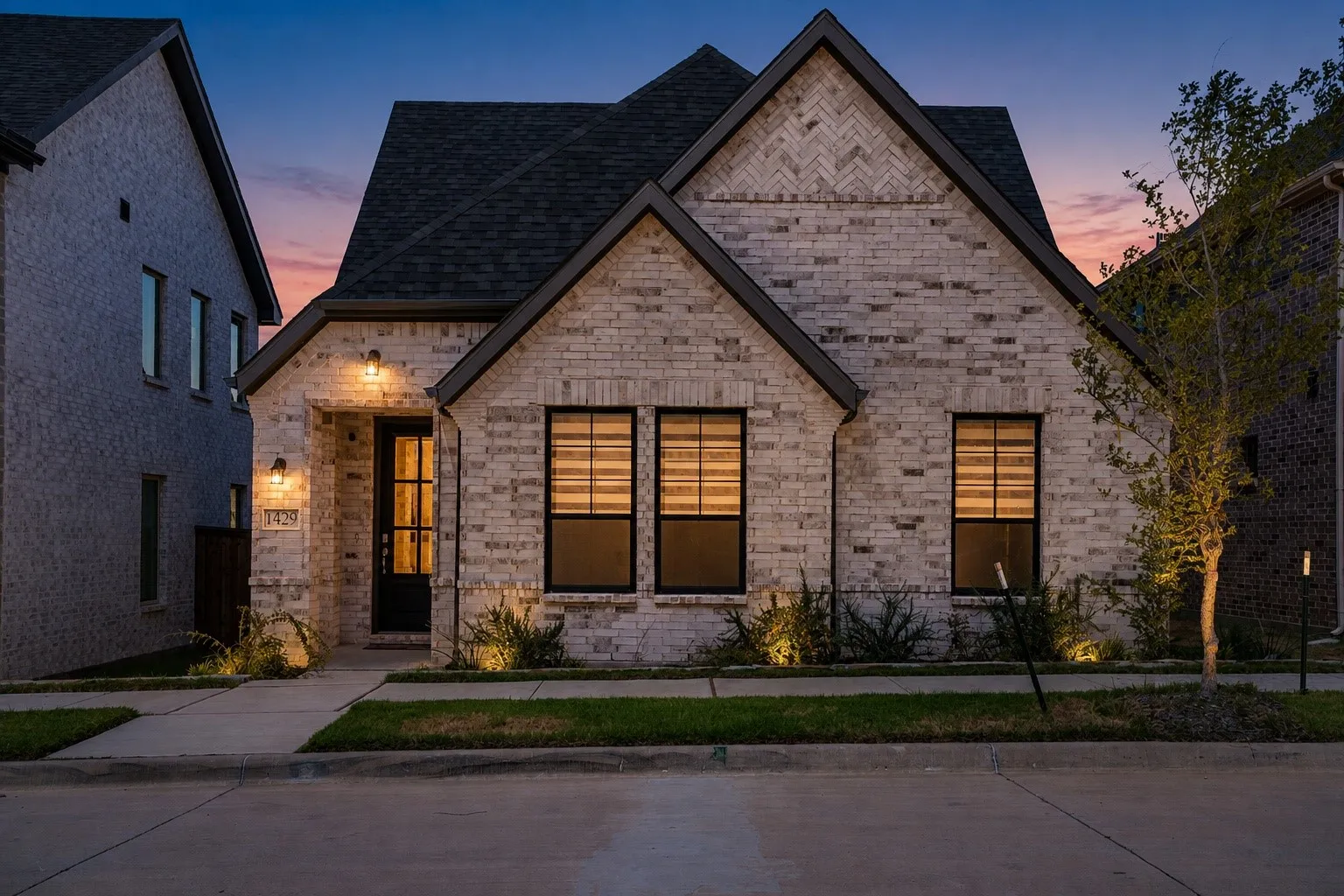 Brick exterior with dark trim accents, gabled roofline, front entry door with glass panels, and well-maintained landscaping with integrated lighting