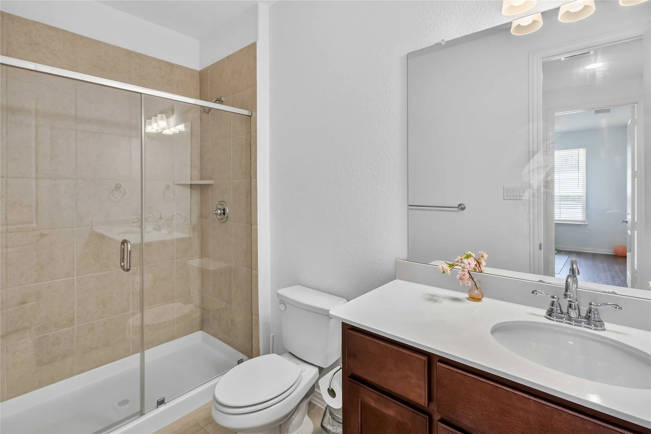 Spa-inspired bathroom featuring a frameless glass-enclosed shower with tile surround, dual-sink vanity with wood-finish cabinetry and white quartz countertop, large mirror with integrated lighting, and chrome faucet fixtures