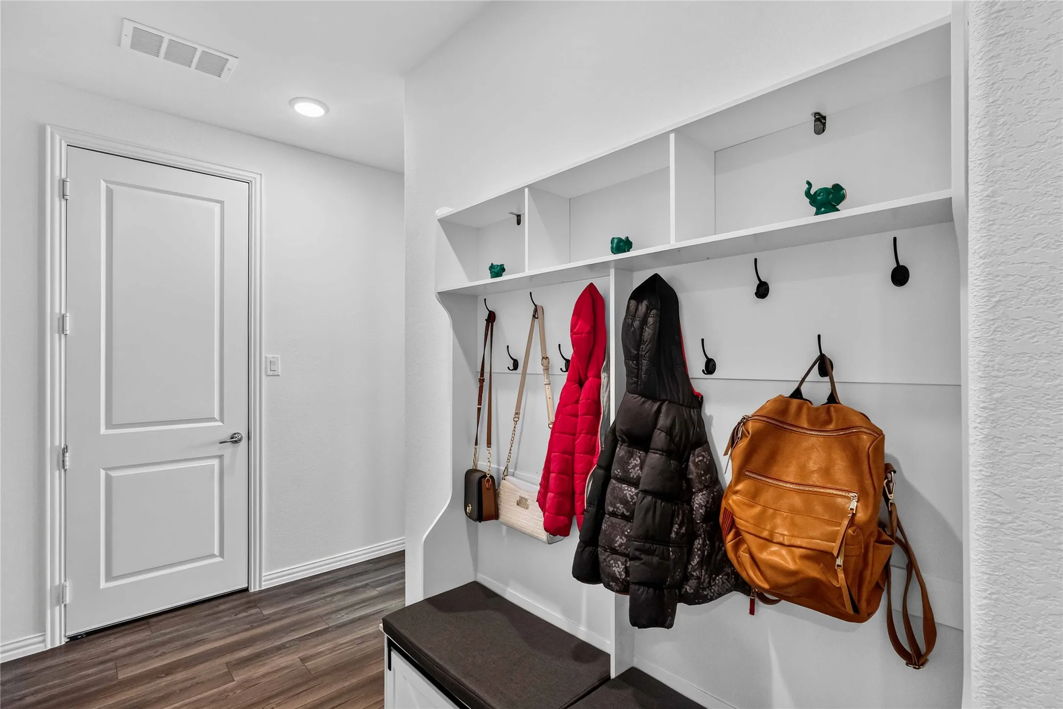 Dedicated mudroom featuring built-in wall-mounted shelving with cubbies, multiple hooks for outerwear storage, wood-finish flooring, and a low-profile bench seating area