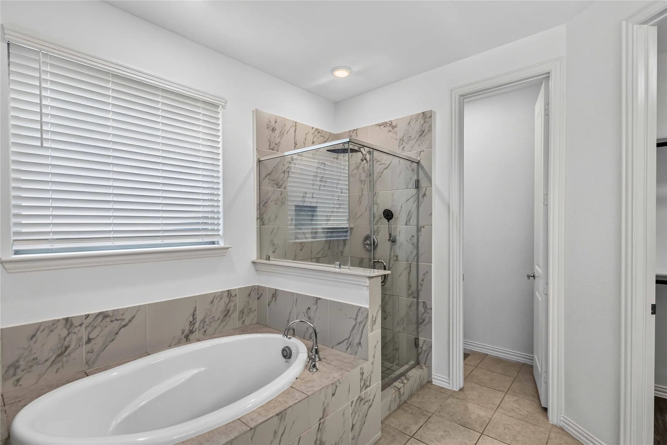 Spa-inspired primary bathroom featuring a soaking tub with stone surround, frameless glass shower enclosure with multi-head fixtures, natural light from horizontal blinds, neutral stone-finish flooring, and a separate water closet