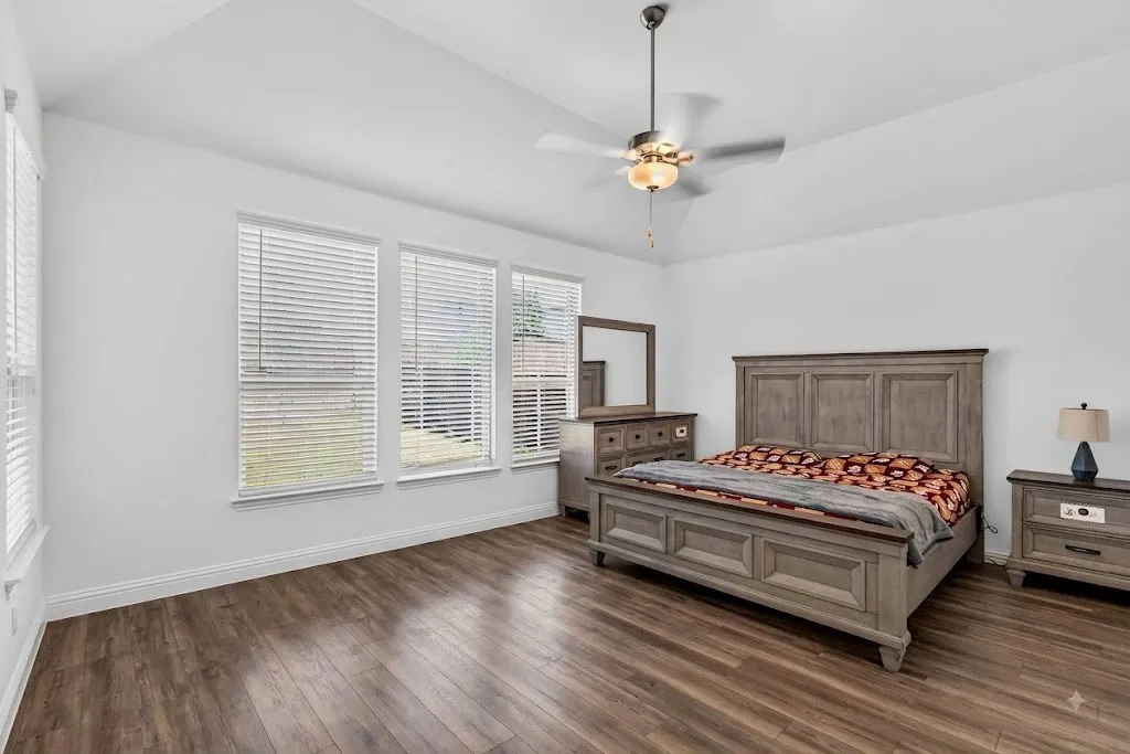 Spacious primary bedroom featuring hardwood flooring, vaulted ceiling with ceiling fan, and expansive multi-pane windows providing natural light