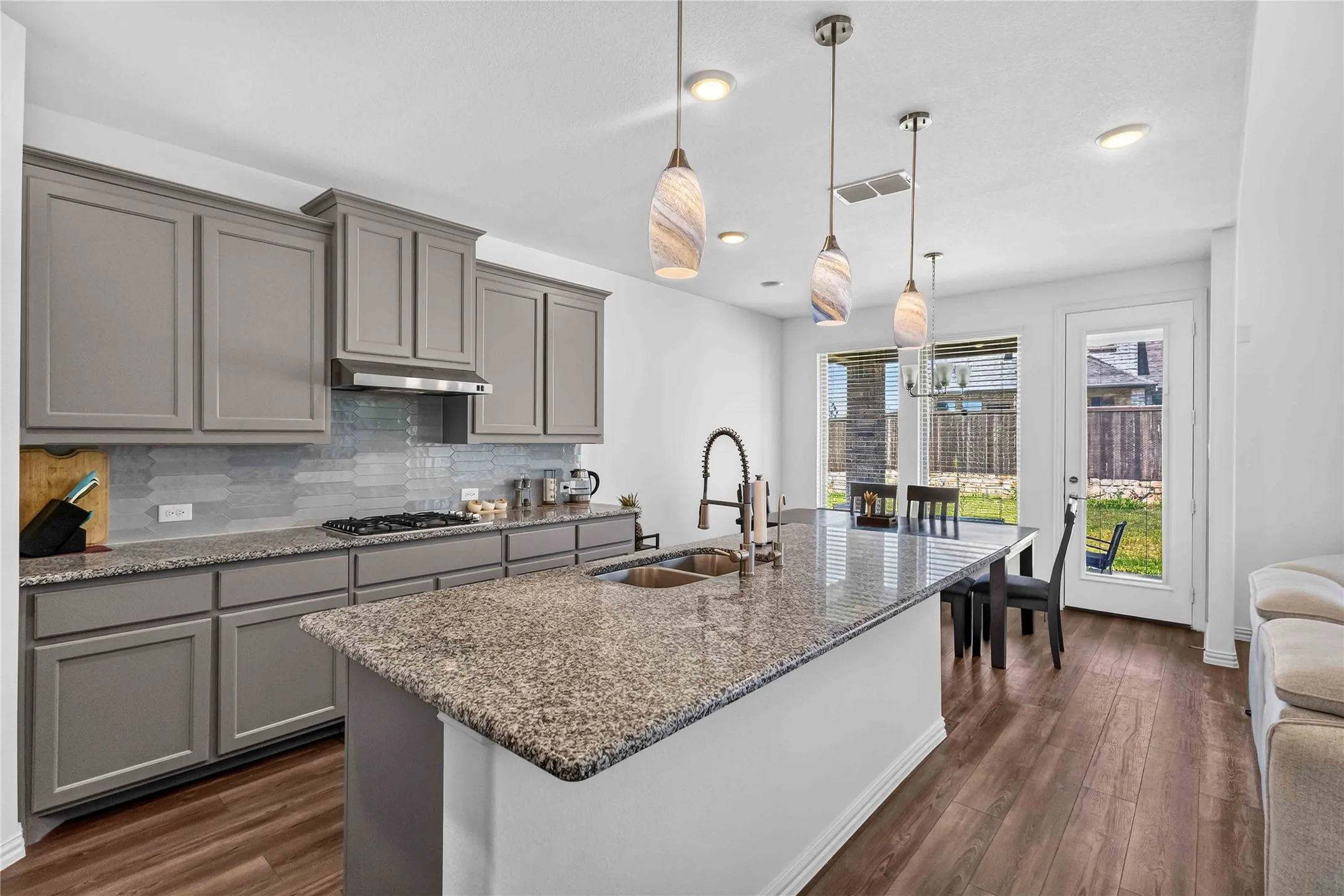 Contemporary kitchen featuring custom cabinetry with refined hardware, granite-topped island with waterfall edge, stainless steel appliances, subway tile backsplash, pendant lighting, and seamless access to the dining and outdoor entertaining spaces via French doors