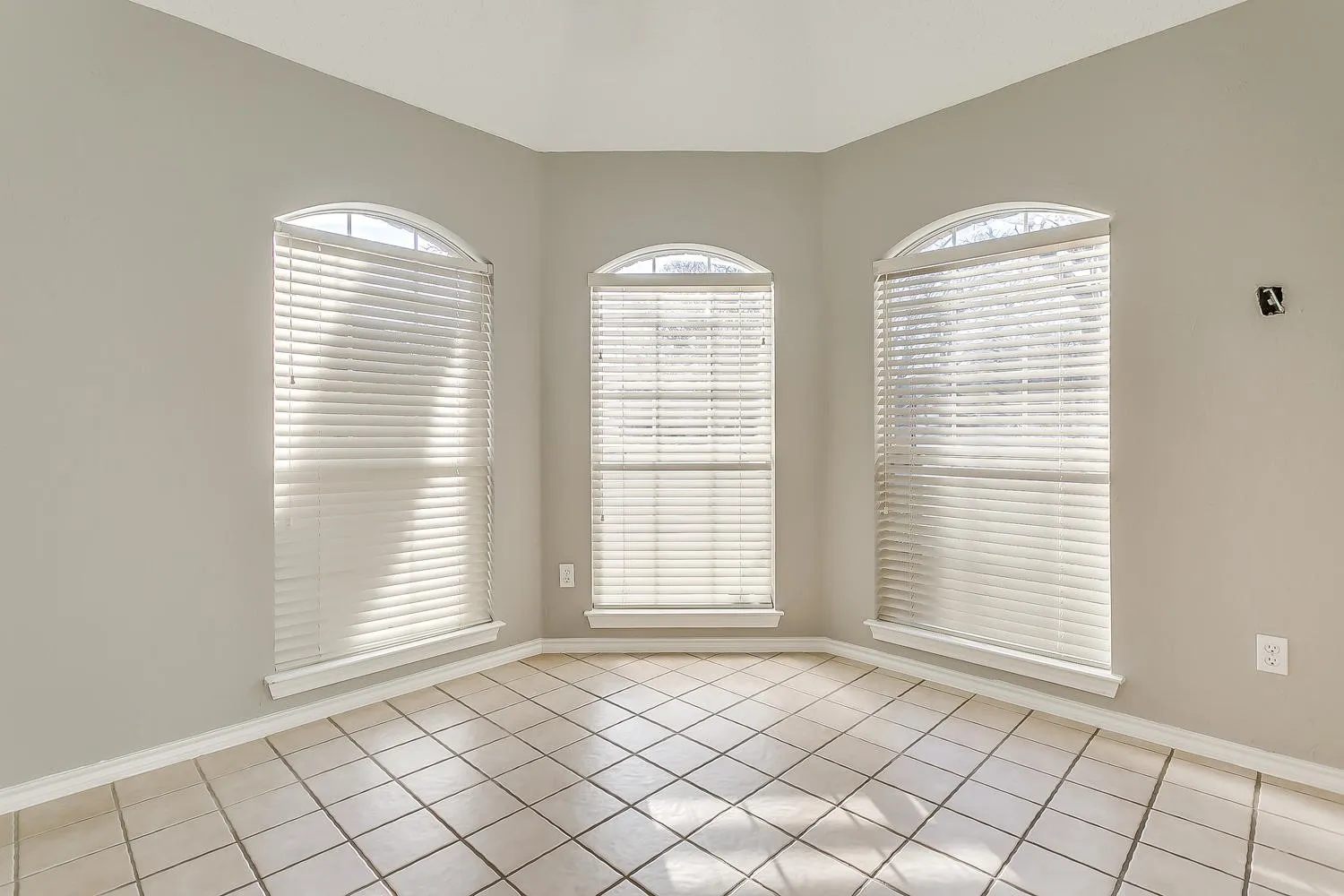 Bright corner room showcasing three arched windows with horizontal blinds, neutral taupe walls, and diagonal ceramic tile flooring