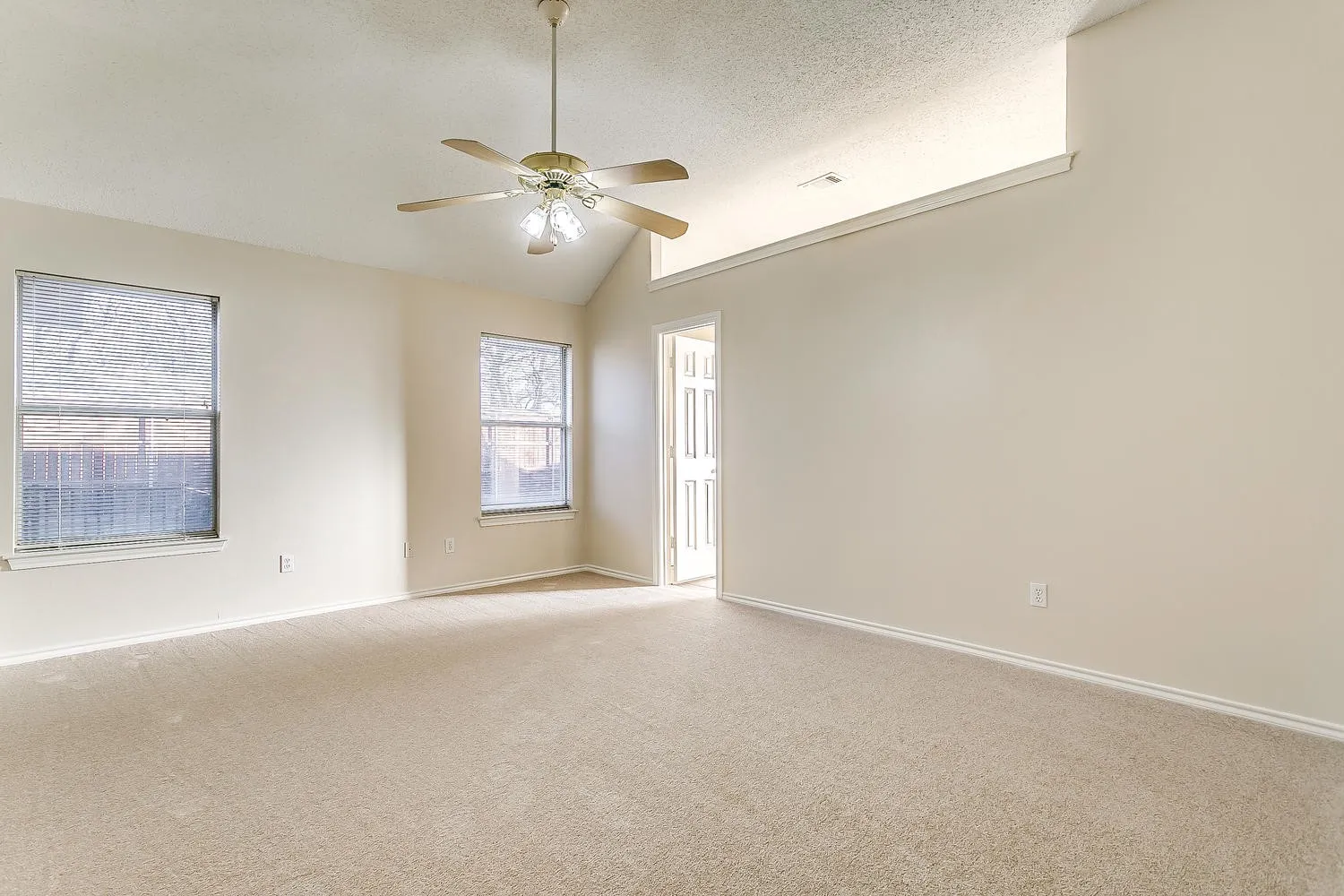 Bright and spacious great room showcasing neutral carpeting, abundant natural light from multiple windows, ceiling fan with integrated lighting, and glass-panel door providing access to outdoor spaces