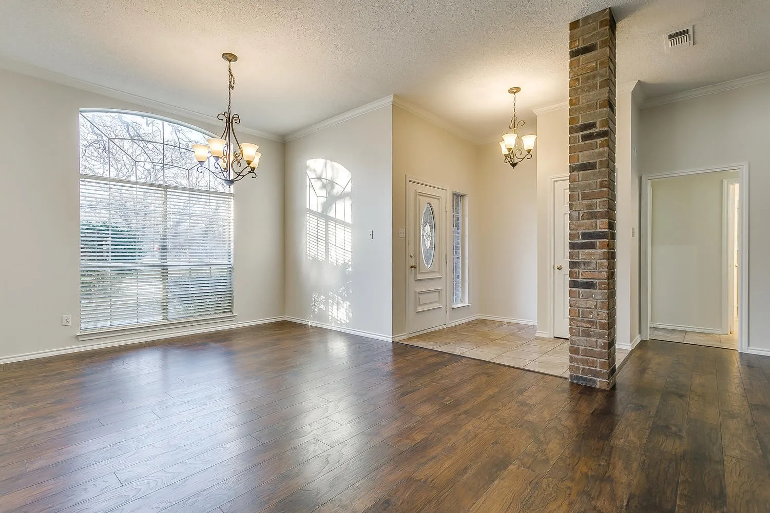 Arched window with blinds, wood-finish flooring, brick accent column, tile entry, and chandelier lighting