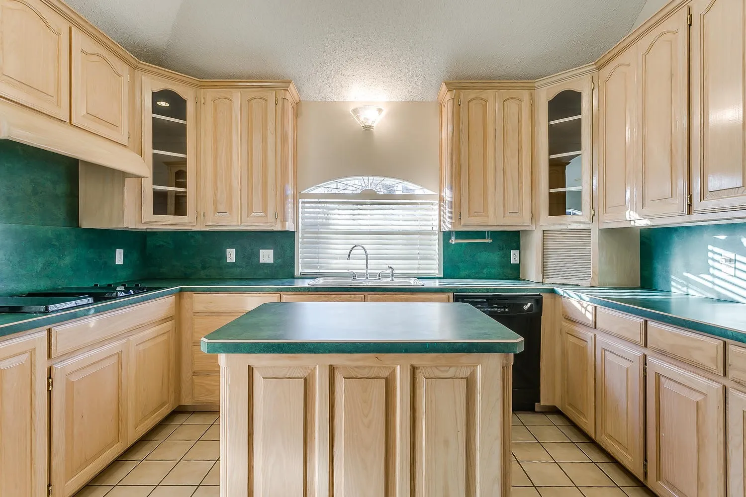 Spacious kitchen featuring natural wood cabinetry with glass-front upper cabinets, a large center island with raised countertop, rich teal tile backsplash, stainless steel appliances, and tile flooring throughout