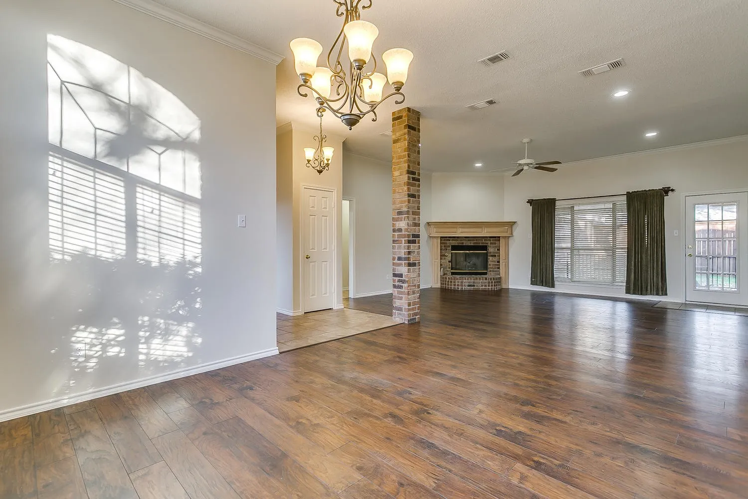 Spacious open living area showcasing gleaming wood-finish flooring throughout, a decorative brass chandelier, and abundant natural light from a large arched window with plantation shutters