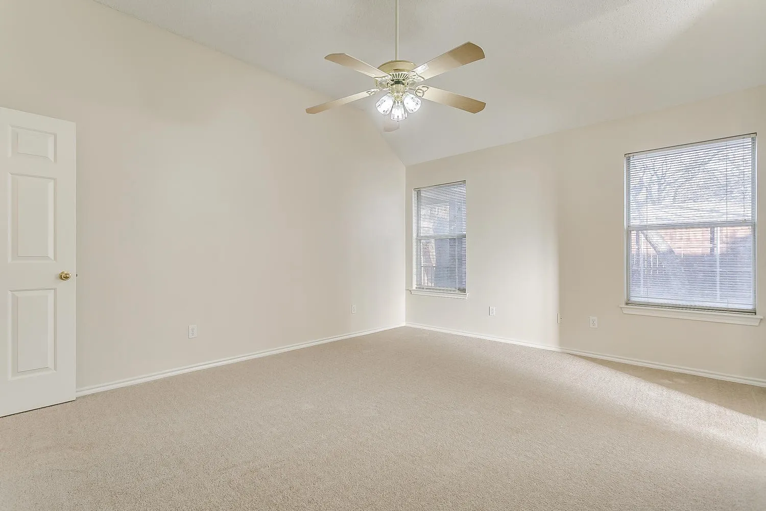 Bright, spacious room featuring neutral carpet flooring, dual windows with natural light, ceiling fan with integrated lighting fixture, and a six-panel entry door