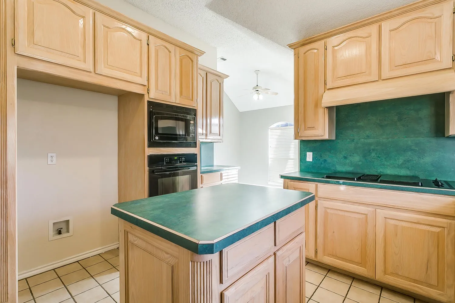 Spacious kitchen featuring warm wood cabinetry, a center island with a teal-edged countertop, stainless steel built-in appliances including a double wall oven and microwave, and a complementary teal tile backsplash