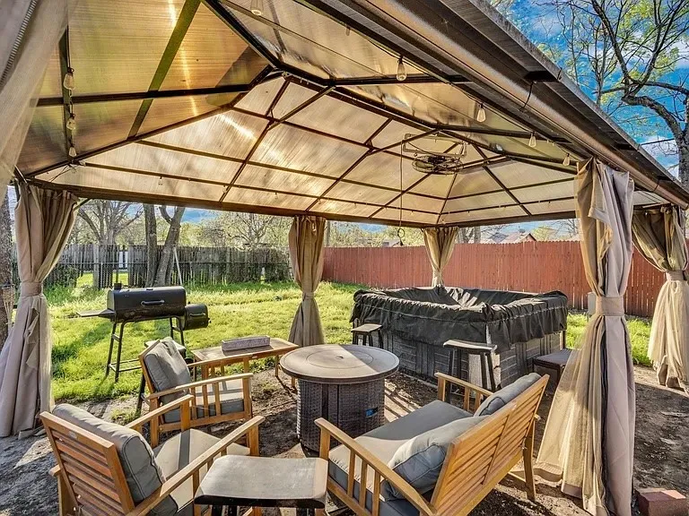 Expansive backyard entertainment space featuring a polycarbonate-roof gazebo with integrated lighting and draped side panels, mature landscaping, manicured lawn, brick fire pit with seating area, and built-in outdoor grill station