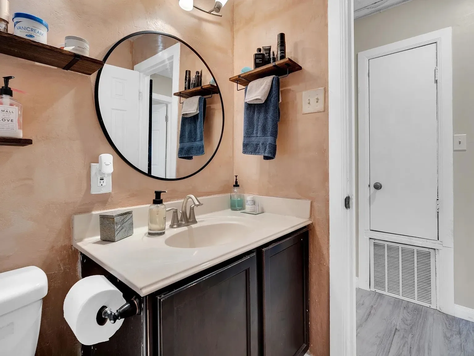 Vanity with a white countertop and integrated sink, dark wood-finish cabinetry, circular wall-mounted mirror, and wall-mounted shelves