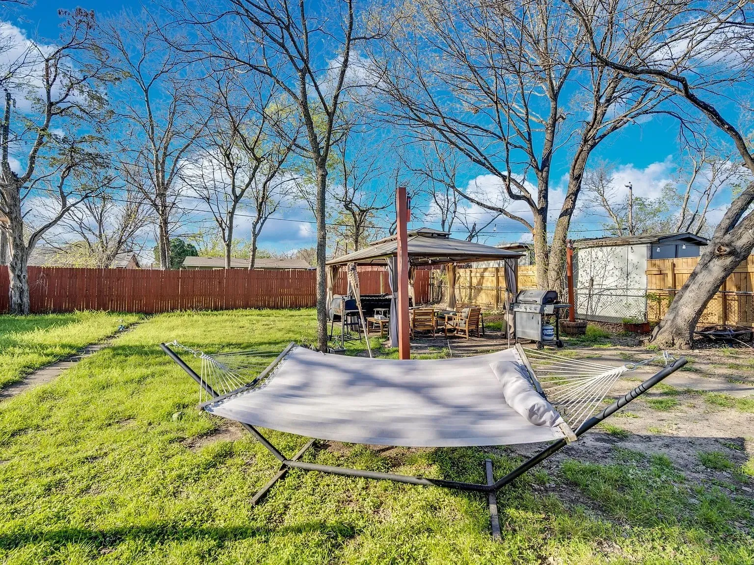 Spacious backyard retreat featuring mature shade trees, a hammock setup, and multiple outbuildings including a covered structure with seating area