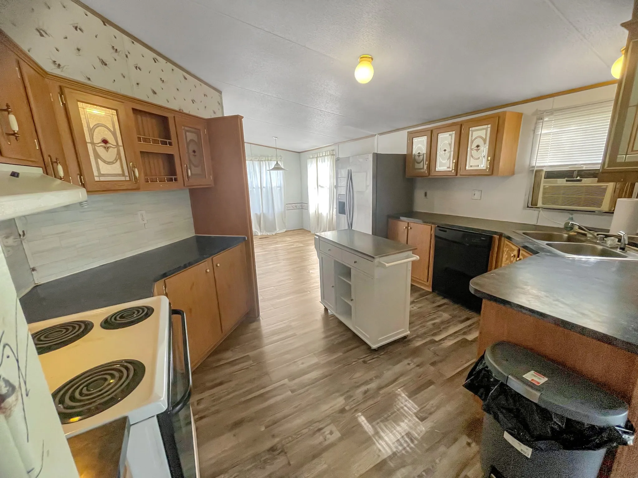 Wood-finish flooring, light wood cabinetry with decorative glass inserts, dark countertops, white kitchen island with storage, and a white electric range