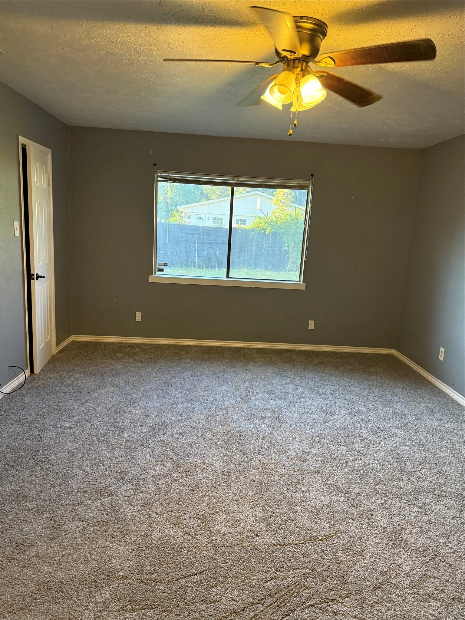 Neutral-toned carpeting, ample natural light from a large window, a ceiling fan with integrated lighting, and a paneled interior door